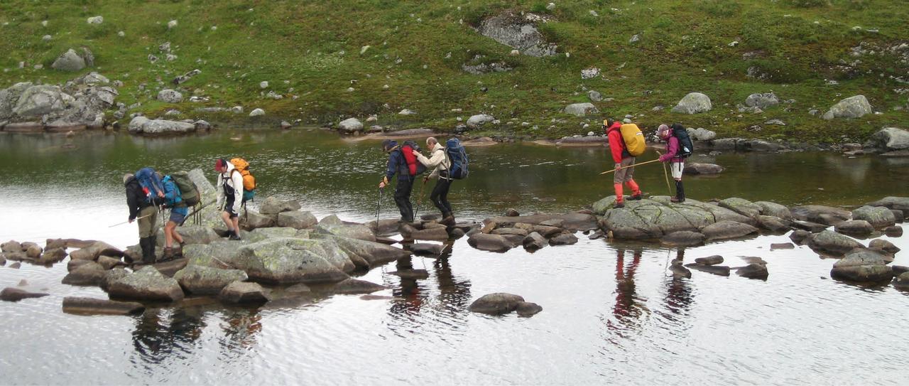 En gruppe krysser en elv. Noen synshemmede holder i sine ledsagere eller i sekken deres, en har kontakt med ledsageren sin ved en ledsagerkjepp